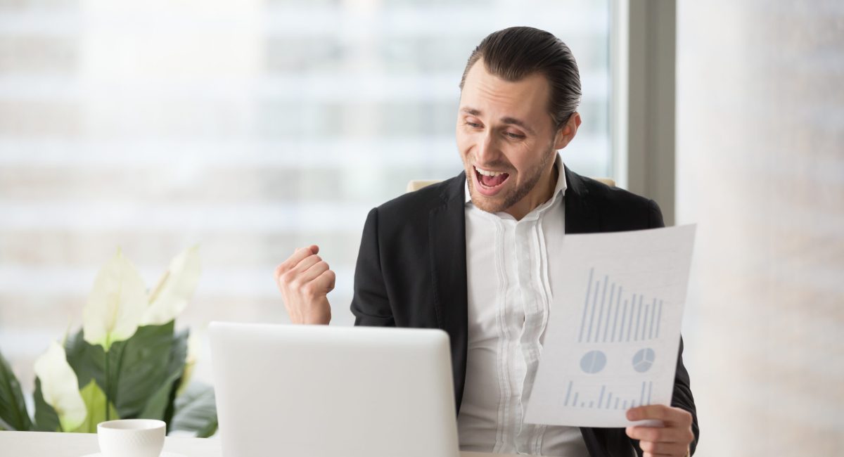 Happy handsome project manager in office celebrates and screams with happiness, looking at laptop screen and holding positive financial report. Great news on stock investment, finance victory concept.